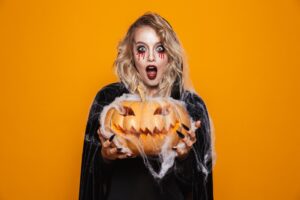 Woman in scary makeup and black Halloween costume holding pumpkin out.